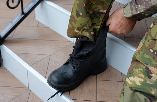 Soldier in Camouflage Uniform Lacing Heavy Black Combat Boots. Close-up of a person in military multicam pants sitting on a step and tying the laces of new, heavy-duty black leather tactical boots