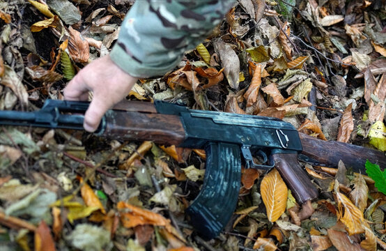 Hand Reaching for Assault Rifle on Autumn Forest Floor. A close-up shot of a person's hand picking up automatic rifle lying among dry autumn leaves
