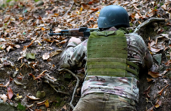 Soldier in Camouflage Lying in Trench with Assault Rifle. Rear view of a soldier wearing a helmet and tactical vest, lying in a defensive position in a forest trench with an assault rifle