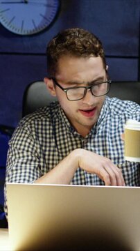 Caucasian man wearing glasses holding coffee cup while using laptop. Male reading information on screen and focusing. Drinking takeaway coffee during work. Sitting at desk in dim room.