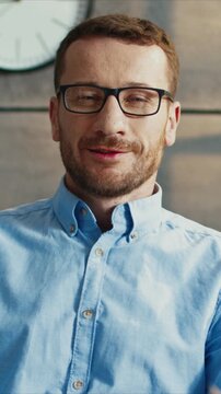 Handsome Caucasian man wearing glasses looking at camera and smiling. Male showing confident expression and relaxed face. Holding steady gaze and feeling satisfied. Sitting in light room.