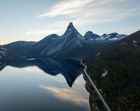 Aerial view of the iconic Stetind mountain peak reflecting in the calm fjord waters with a winding coastal road in Narvik Municipality, Nordland, Norway.