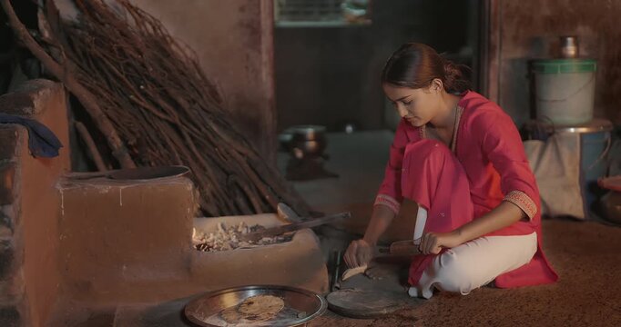 Indian gen z young chef woman sitting do work make fresh roti use mud clay stove indoor house Asian teen age girl cook casual food on open fire flam kitchen rural area home