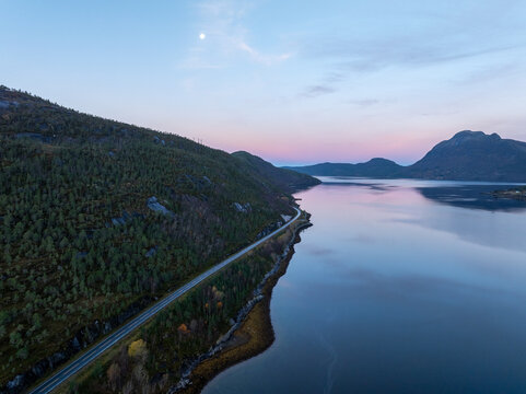 Aerial view of the calm waters of a fjord reflecting a pink twilight sky with a bright moon above a coastal road and forested hills in Narvik Municipality, Nordland, Norway.