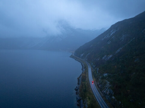 Aerial view of the iconic Stetind mountain shrouded in mist above a winding coastal road with a car driving along the dark fjord in Narvik Municipality, Nordland, Norway.