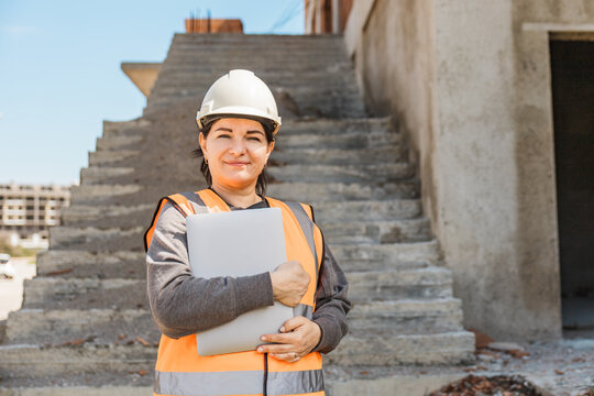 woman stands on construction stairs wearing a hard hat and safety vest, holding a laptop. The construction site is visible in the background.