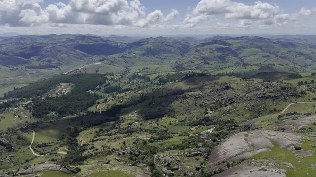 Drone flies left high over rolling green hills under shadows of clouds on a sunny day at Sibebe Rock near Mbabane, Eswatini