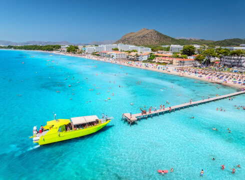 Aerial drone view of Alcudia white sandy beach and yellow tour boat, swimming people, sunbathing, pier, hotels. Mallorca, Balearic Islands, Spain. Top view of yacht, blue sea, sandy beach, mountain