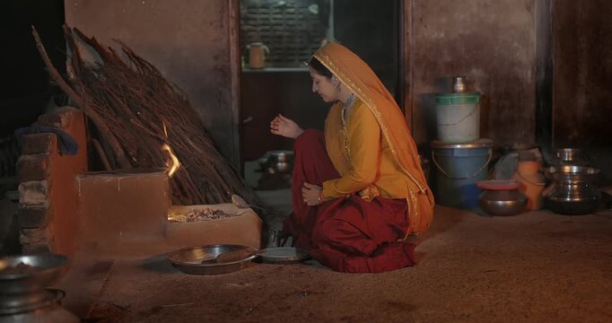 Indian mature adult chef woman mom wear saree sitting do work cook food local dark room rural area. Female lady use metal chimta make fresh roti chapati on open fire flam clay stove home place