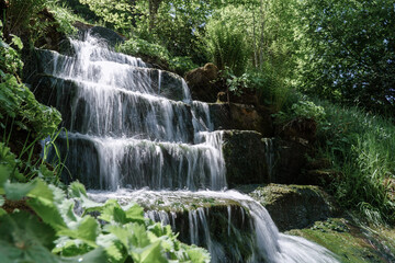 Obraz premium Cascading forest waterfall flowing smoothly over stone steps in long exposure with silky water motion and soft misty atmosphere in summer sunlight