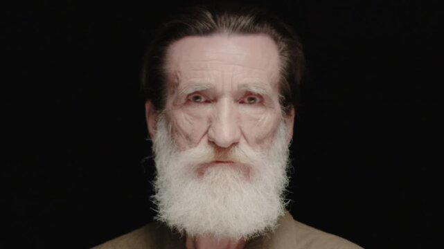 Close portrait of senior Caucasian man with long beard looking at camera. Male showing calm expression and wrinkles. Holding steady gaze. Standing against dark background in studio.