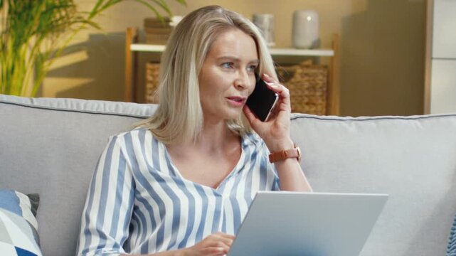 Caucasian woman holding smartphone while using laptop on sofa. Female talking with client and checking screen. Discussing details and reviewing data. Working from home in light room.