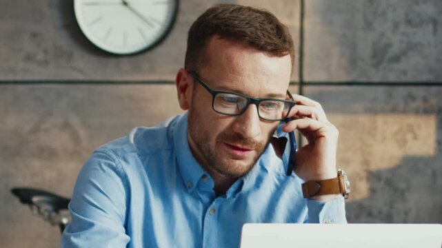 Caucasian man in glasses talking on smartphone while working on laptop. Male checking data on screen. Discussing details with client. Sitting at desk in light room. Having working online communication