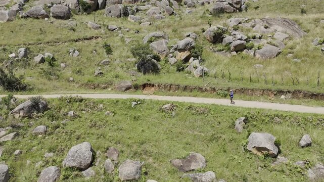 Drone flies forward as hiker crosses the frame on a trail and drone rises up to reveal the top of trail on a sunny day at Sibebe Rock near Mbabane, Eswatini