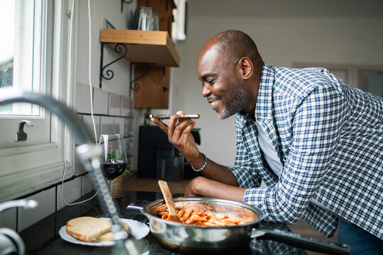 Middle-aged man cooking pasta in home kitchen using smartphone voice assistant