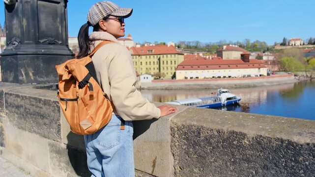 A traveler enjoys the stunning views of Prague from a riverside location. The warm sunshine illuminates the historic buildings across the water, creating a picturesque holiday atmosphere.