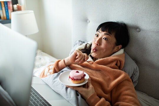 Woman relaxing in bed with dessert and laptop at home