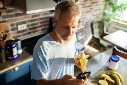 Middle aged man eating banana while checking smartphone in home kitchen