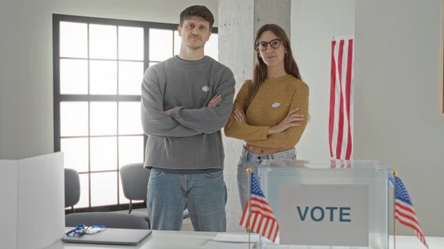 Two young voters with crossed arms beside a clear ballot box and american flag in a polling building; civic duty pride.