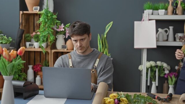 Man at laptop looks up as woman florist holds bouquet with both hands and gestures in a flower studio; collaboration focus.