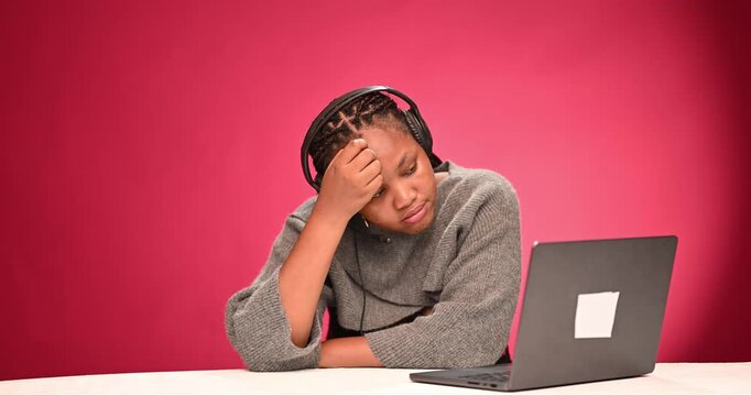 Young woman sitting at desk using laptop appearing tired and frustrated while working or studying online against pink studio background