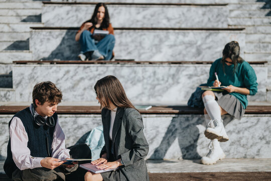 A group of students study and interact on marble campus steps with notebooks. Two students discuss notes while others study alone on the outdoor benches.