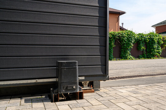 black sliding gate with an automatic drive mechanism, mounted on a paved area. Behind it a wooden fence covered with green ivy, adjacent to brick houses under a clear sky