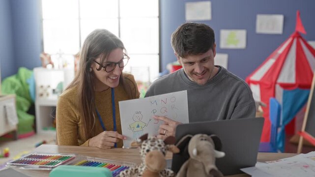 Young woman teacher and man review child's drawing by a laptop, woman pointing at sketch in a preschool classroom building; joyful teamwork.