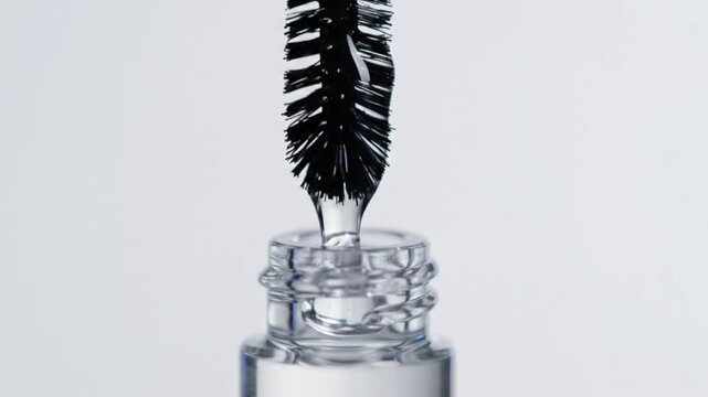 Close-up of a mascara brush being removed from its container, dripping black liquid against a white backdrop.