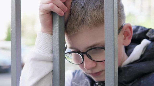 A teenage boy stands behind a metal fence, looking upset and isolated. The scene conveys sadness and bullying at school, highlighting emotional struggle and vulnerability. 