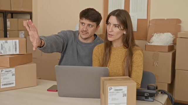 Young man raises hand palm toward woman over laptop and stacked parcels in a small business building; teamwork focus.