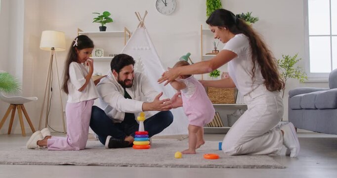 First steps of baby with help of mother, happy parents support barefoot toddler walking at home. Family enjoying toddler milestones together, smiling woman holding hands of child in living room