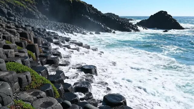 Powerful ocean waves crashing against hexagonal basalt columns on a sunny day with blue water and sky