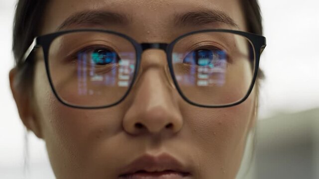 Extreme close-up video of a professional's eyes in black-rimmed glasses reflecting cool digital data, dollying out to reveal their focused expression in a minimalist workspace with creamy bokeh.