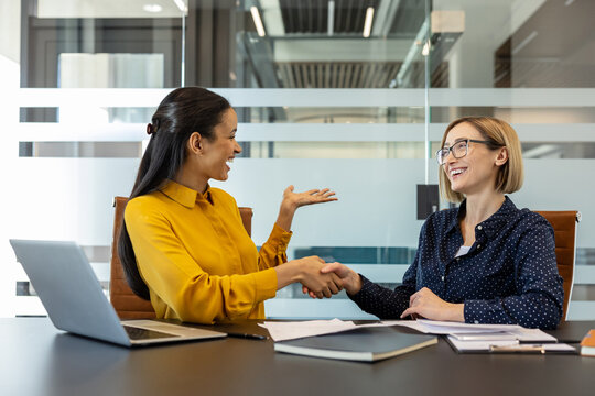 Two businesswomen are smiling and shaking hands across an office desk with a laptop and documents, symbolizing a successful partnership, collaboration, and agreement