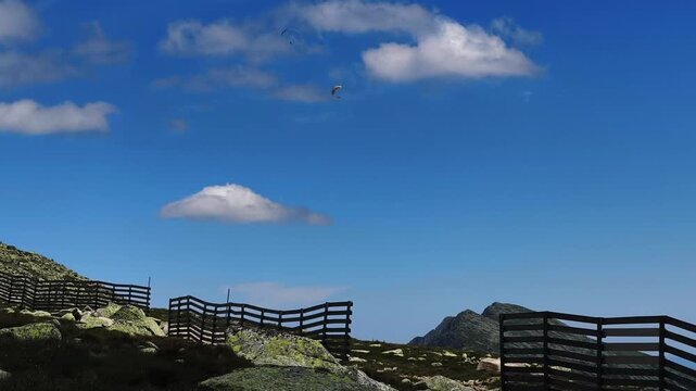 Stunning mountain landscape featuring a wooden fence along a rocky trail. Two paragliders are flying high in the bright blue sky with white clouds in Demanovska Dolina, Slovakia