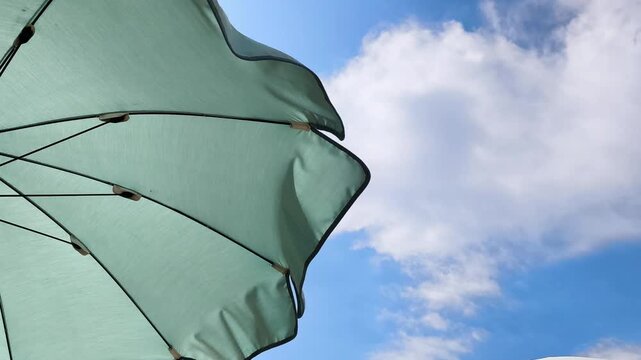 Low angle view of a light green beach umbrella blowing in the summer breeze. White clouds are drifting across the bright blue sky in Pineta, Lignano Sabbiadoro, Italy