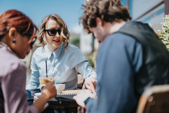 Business coworkers collaborate outdoors at a cafe, reviewing documents and notes. Three colleagues discuss work over coffee during an informal outdoor meeting.