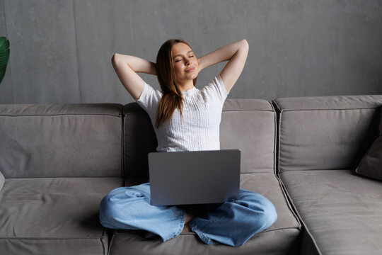 Young woman is stretching her arms while sitting on a sofa working remotely on her laptop in her cozy living room
