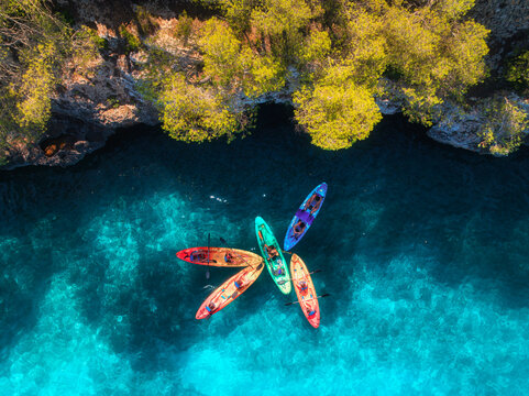 Aerial drone view of colorful kayaks with people in crystal clear turquoise sea near rocky coast and green trees. Summer outdoor adventure in Mallorca, Balearic Islands, Spain. Top down view of canoes