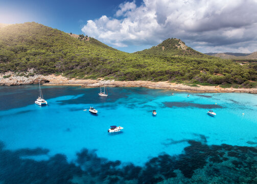 Aerial view of luxury yachts and speed boats on blue sea, beach, mountain with green forest, sky with clouds in summer. Top drone view of Cala Agulla, Mallorca, Spain, Balearic Islands. Blue lagoon