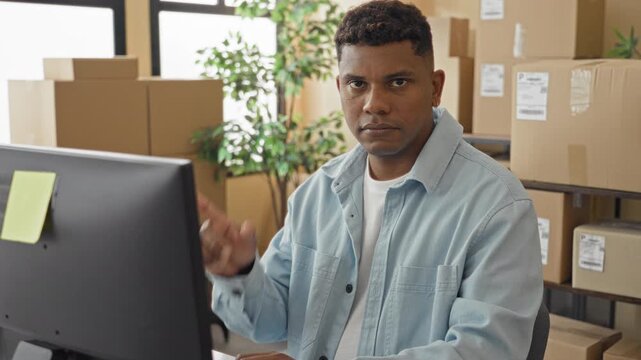 Man pointing finger at computer while typing beside stacked shipping boxes and parcels in a packing building; focused.