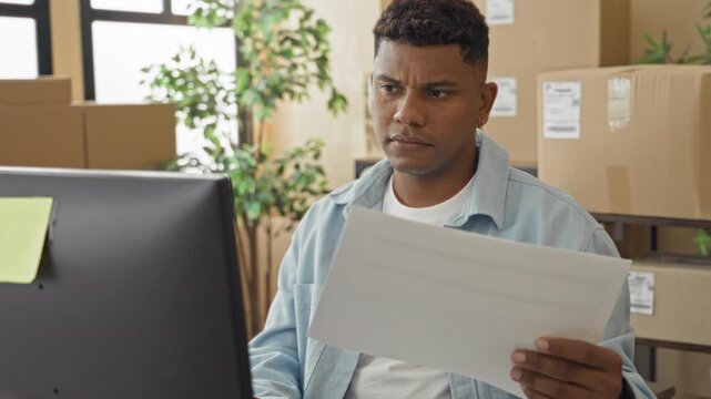 Man frowning while reading a delivery document at a computer amid stacked packages in a building; frustration paperwork.