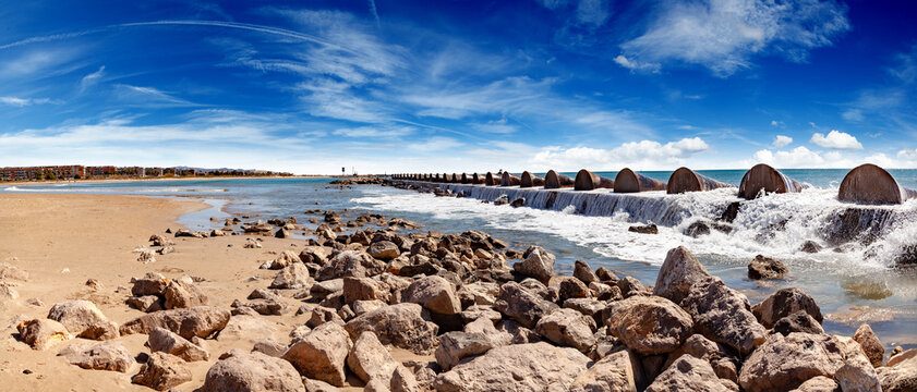 Playa de Cubelles, Catalu&ntilde;a, Espa&ntilde;a, costa mediterr&aacute;nea. Playa rocosa y arenosa con rompeolas, olas rompiendo, mar azul en calma y edificios costeros bajo un cielo brillante.