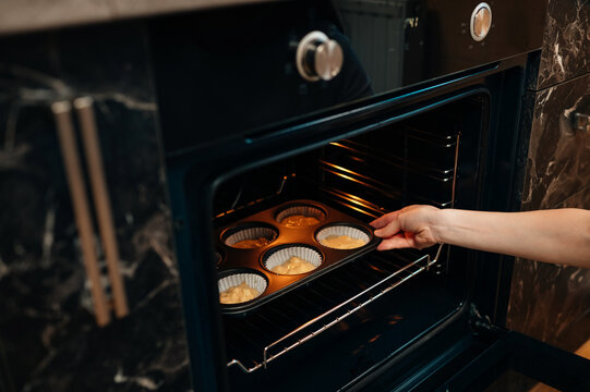 Home Baker Placing Muffin Tray In Oven, Warm Kitchen Light, Dark Marble Countertops, Brass Knobs, Closeup