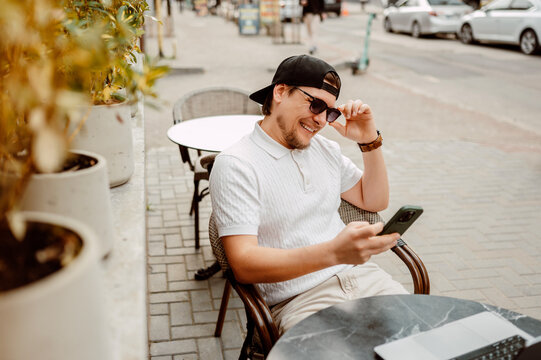 Man Laughing At Phone While Seated, Outdoor Cafe Table, Laptop Closed Nearby, Coffee Cup On Table, Relaxed