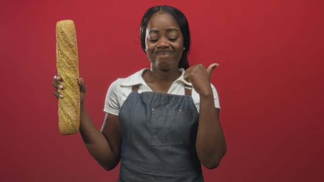 Woman baker wearing denim apron holds long baguette in left hand and points thumb to shoulder while gazing at camera in studio; pride craft skill.