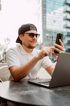 Man Using Phone And Laptop At Outdoor Cafe, Casual Cap And Sunglasses, Focused On Mobile Productivity