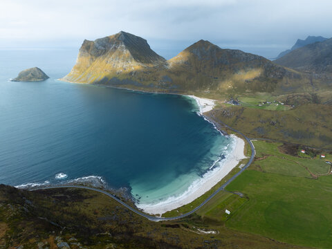 Aerial view of the turquoise waters and white sands of Haukland Beach surrounded by steep mountains and green fields in the Lofoten islands, Nordland, Norway.