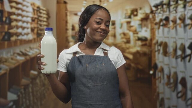 Woman holding milk bottle in hand, smiling and wearing denim apron amid shelves of shoes in a shoe shop building; pride.